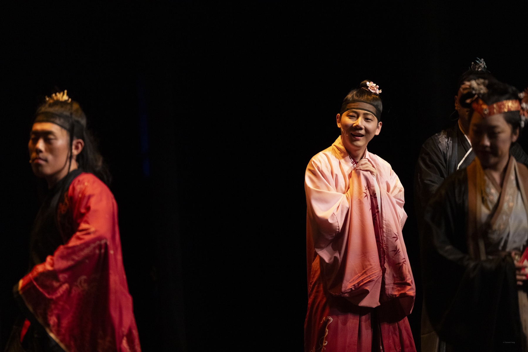 Hua performs in a pink period costume under a spotlight with the ensemble around him.
