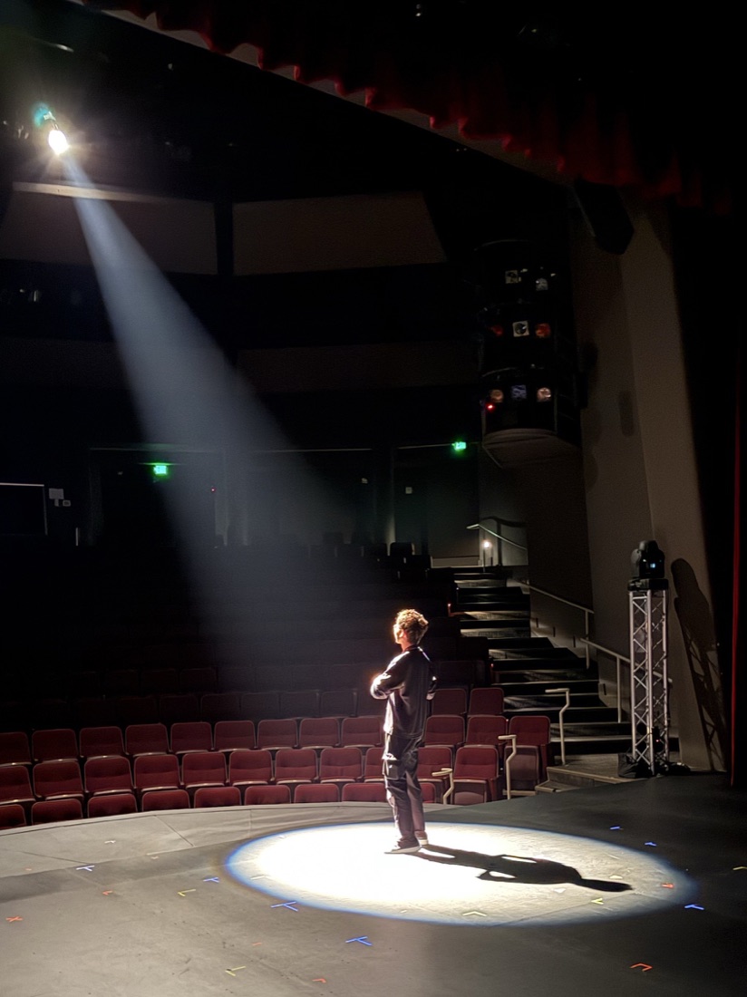 A performer stands alone in a spotlight while preparing on an empty theatre stage.